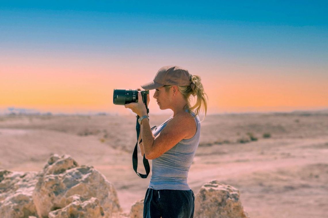 Side view of serious female photographer in casual clothes and cap standing with photo camera and taking pictures near rough rocks in sandy valley under cloudless sky in daytime