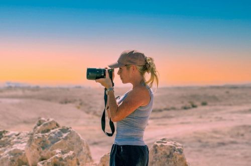 Side view of serious female photographer in casual clothes and cap standing with photo camera and taking pictures near rough rocks in sandy valley under cloudless sky in daytime