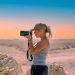 Side view of serious female photographer in casual clothes and cap standing with photo camera and taking pictures near rough rocks in sandy valley under cloudless sky in daytime