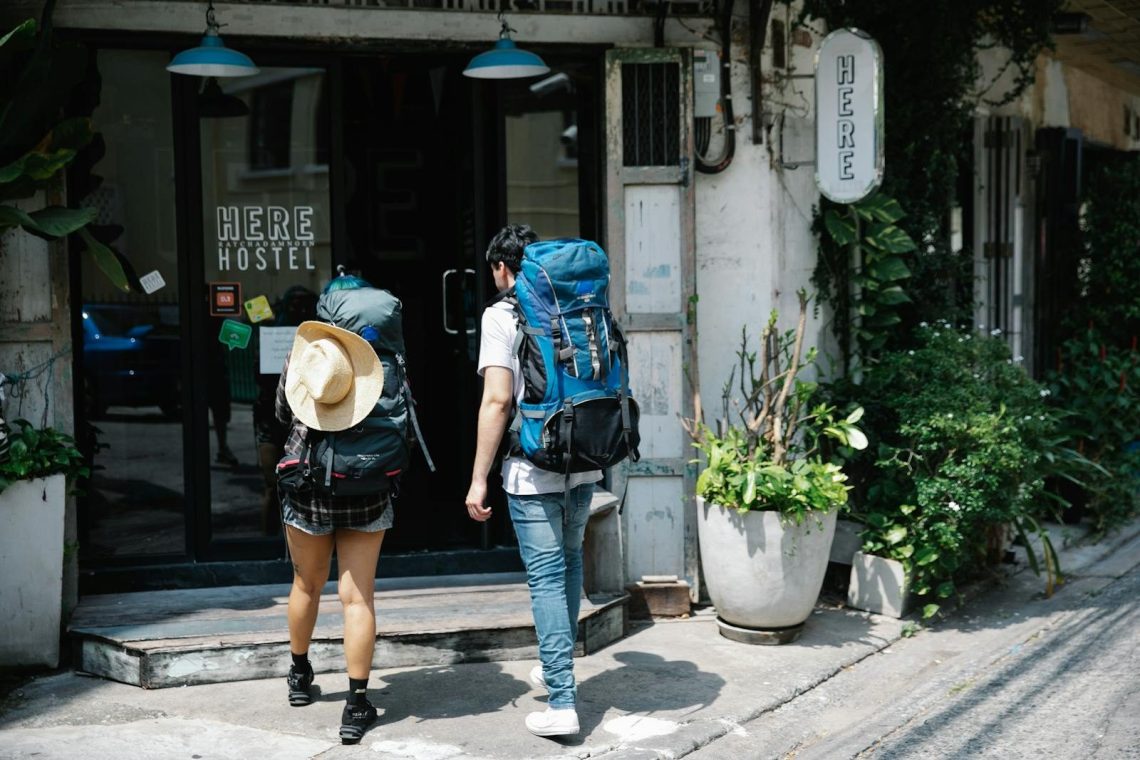 Back view of anonymous couple of tourists with rucksacks walking on street and entering hostel door during trip